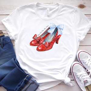 May include: White t-shirt featuring a watercolor illustration of red glittery high-heeled shoes with bows. The shoes have a silver gem and a blue gingham bow. Text on the shirt reads "There's No Place Like Home".