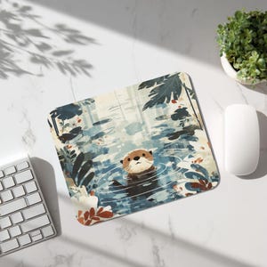Puede incluir: Alfombrilla de ratón rectangular con una ilustración de una nutria nadando en un estanque. La obra de arte incluye agua azul, follaje verde y una nutria marrón claro. La alfombrilla está sobre una superficie blanca con teclado, ratón y planta.