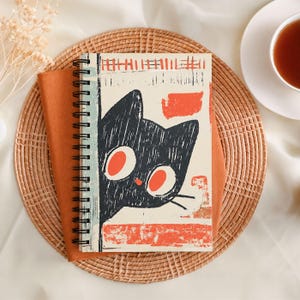Peut inclure: Un carnet &agrave; spirales avec une illustration de chat noir aux grands yeux orange et au nez rouge. Le carnet repose sur un tissu orange et un dessous de plat en osier, avec une tasse de th&eacute; en arri&egrave;re-plan.