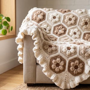 May include: A handmade crochet blanket featuring a repeating hexagonal pattern with raised, textured floral motifs in shades of cream, beige, and light brown. The blanket is finished with a wide, ruffled border in a solid cream yarn color.