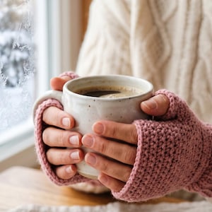 May include: A person holding a white ceramic mug filled with coffee, wearing pink crocheted fingerless gloves. The background includes a window with a frosted winter scene and a cream-colored cable-knit sweater.