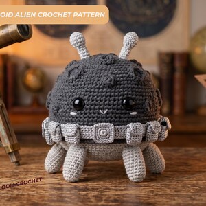 Puede incluir: Una figura alienígena de ganchillo gris oscuro, redonda y texturizada, con dos antenas, grandes ojos negros y un cinturón utilitario gris claro con múltiples bolsillos. La figura tiene cuatro patas gris claro e incluye el texto ASTEROID ALIEN CROCHET PATTERN.