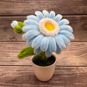 May include: A handcrafted, fluffy blue daisy flower with a white and yellow centre, presented in a small white pot. The flower has green leaves and stem. The background is a wooden surface.