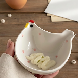 May include: A white ceramic chicken-shaped bowl with a red comb and yellow beak, decorated with pink polka dots. The bowl contains a white condiment. A small whisk with a wooden handle is next to the bowl. The bowl is held by a person.