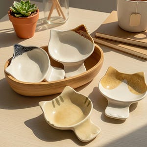 Puede incluir: Un conjunto de cuencos de cerámica con forma de gato en varios colores, incluyendo blanco, beige y marrón. Los cuencos están dispuestos en un cuenco de madera y sobre una superficie clara. También se ve una taza de té y una pequeña planta en maceta.