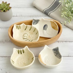 May include: A collection of four ceramic cat-shaped dishes. Two are presented in an oval wooden tray. The dishes are white with painted details in grey, brown, and green. The dishes are designed to resemble cats.