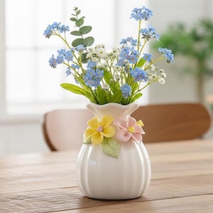 Pode incluir: Um vaso de cerâmica branca com corpo canelado e borda ondulada, adornado com detalhes florais amarelos e rosa. O vaso contém um buquê de flores azuis e brancas com folhagem verde, criando um arranjo fresco e primaveril.