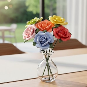 May include: A clear glass vase holding a bouquet of colorful ceramic roses. The roses are in shades of pink, orange, yellow, red, blue, and green. The vase sits on a wooden table with a white tablecloth.