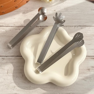 Puede incluir: Un conjunto de pinzas de servir de acero inoxidable con diseños únicos, incluyendo extremos en forma de bola, agarres en forma de mano y puntas en forma de pétalo. Las pinzas están dispuestas sobre un plato crema en forma de flor.