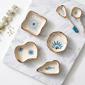 Könnte beinhalten: Eine Sammlung von cremefarbenen Keramik-Servierschalen und -löffeln mit blauen floralen und geometrischen Mustern. Die Schalen haben unregelmäßige Formen und braune Ränder. Zwei Löffel haben blaue Griffe und florale Muster.