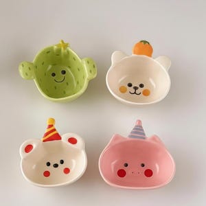 以下が含まれることがあります： サボテン、クマ、ネコ、パーティーハットをかぶったクマの形をした4つのセラミックボウル。サボテンのボウルは緑色、クマのボウルはオフホワイト、ネコのボウルはピンク色です。各ボウルには笑顔があり、軽食や少量の食事を提供するように設計されています。