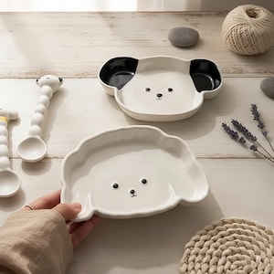 Könnte beinhalten: Weiße Keramikplatten in Hundeform mit schwarzen Akzenten und Gesichtsmerkmalen. Die Teller stehen auf einer hellen Holzoberfläche, zusammen mit einem weißen Löffel mit Hundekopf-Design. Die Teller sind zum Servieren von Speisen bestimmt.