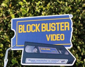 May include: A sticker set featuring a blue and yellow Blockbuster Video logo and a VHS tape. The tape has the words "BE KIND REWIND" and "Please remember to rewind!" printed on it. The background is green foliage.