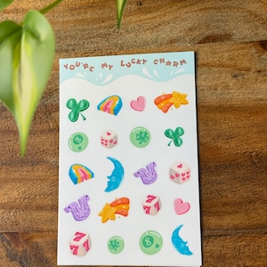 May include: A sheet of stickers with the text "You're my lucky charm." The stickers feature various lucky symbols, including shamrocks, rainbows, hearts, shooting stars, dice, and a crescent moon. The stickers are in various colors, including green, pink, orange, and blue.