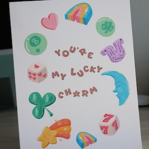 May include: A white greeting card with colorful illustrations of lucky charms, including a heart, dice, and a four-leaf clover. The text "You're My Lucky Charm" is written in brown letters. The card is perfect for sending to a loved one.