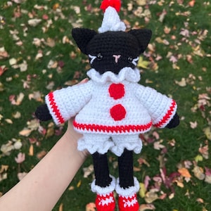 May include: A handmade crochet cat doll wearing a white outfit with red accents. The cat has a black head and legs, a white ruffled collar, and a red pom-pom on its hat. The doll is approximately 8 inches tall.