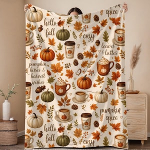 May include: A cream-colored throw blanket featuring a repeating pattern of orange and green pumpkins, brown autumn leaves, pinecones, and mugs of coffee with text including hello fall, pumpkin kisses, harvest wishes, cozy, and pumpkin spice.