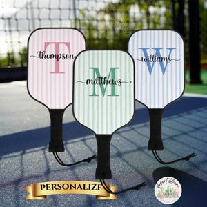 Könnte beinhalten: Drei personalisierte Pickleball-Schläger mit gestreiften Hintergründen in Rosa, Mintgrün und Blau. Jeder Schläger hat einen großen Anfangsbuchstaben und einen Nachnamen. Die Schläger haben schwarze Griffe und sind für die Personalisierung bestimmt.