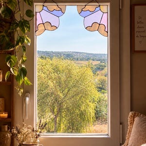 May include: Two stained glass window hangings shaped like stylised clouds, featuring pastel purple, blue, and cream glass panels with dark metal soldering, suspended by thin chains from the top corners of a white window frame overlooking a landscape.