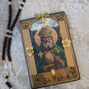 Op de afbeelding: Een ingelijste afbeelding van St. Patrick met een gouden kruis en bloemaccenten. De afbeelding is omgeven door een gouden rand en de woorden "St. Patrick". Een rozenkrans met donkerbruine kralen en een zilveren kruis staat links.