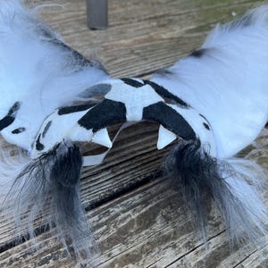 Pode incluir: Um par de orelhas peludas brancas e pretas com um centro manchado em preto e branco. As orelhas t&ecirc;m detalhes de penas pretas e dois dentes brancos. O acess&oacute;rio &eacute; provavelmente para cosplay ou fantasia.