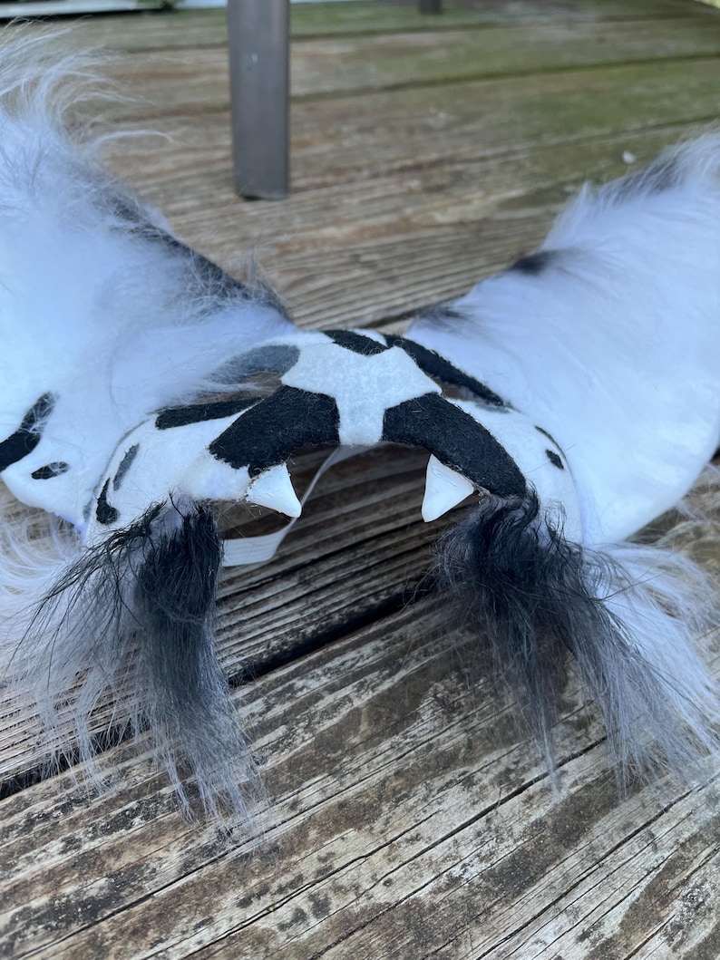 Pode incluir: Um par de orelhas peludas brancas e pretas com um centro manchado em preto e branco. As orelhas t&ecirc;m detalhes de penas pretas e dois dentes brancos. O acess&oacute;rio &eacute; provavelmente para cosplay ou fantasia.