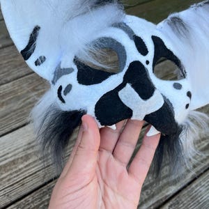 Pode incluir: Uma m&aacute;scara de animal com manchas brancas e pretas com orelhas grandes e acabamento peludo. A m&aacute;scara tem manchas pretas e um nariz preto, com dentes brancos. A m&aacute;scara &eacute; segurada por uma m&atilde;o.