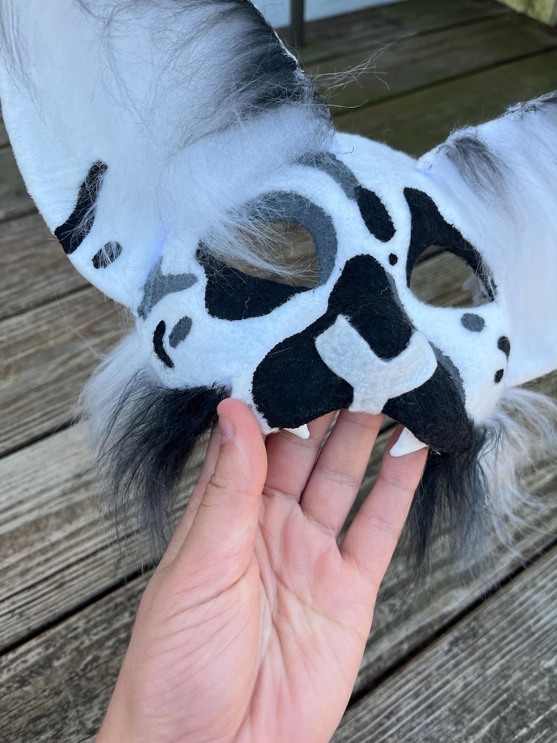 Pode incluir: Uma m&aacute;scara de animal com manchas brancas e pretas com orelhas grandes e acabamento peludo. A m&aacute;scara tem manchas pretas e um nariz preto, com dentes brancos. A m&aacute;scara &eacute; segurada por uma m&atilde;o.