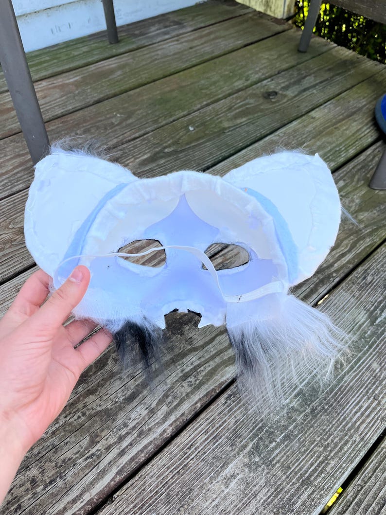Pode incluir: Uma m&aacute;scara de pele branca e azul claro com orelhas grandes e detalhes em preto e branco. A m&aacute;scara &eacute; segurada por uma m&atilde;o e est&aacute; sobre uma superf&iacute;cie de madeira. A m&aacute;scara tem buracos para os olhos e uma al&ccedil;a.