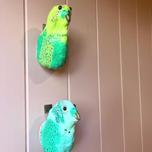 May include: Two wall-mounted bird sculptures crafted with a textured finish. The top bird is bright lime green with dark green wing markings and a gold beak. The bottom bird is turquoise with dark teal wing markings and a gold beak.