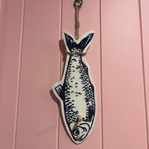 May include: A white wall-mounted fish-shaped hook with dark blue hand-painted scale and fin details. The decorative piece features a natural jute rope loop at the top for hanging and a small wooden peg protruding from the lower body section.