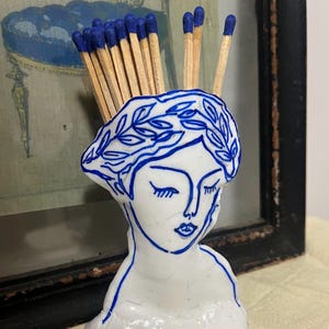 May include: A white ceramic match holder shaped like a woman's head, decorated with blue line art. It contains wooden matches with blue tips. The background features a framed print with blue details.