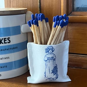 Könnte beinhalten: Ein weißer Keramik-Streichholzhalter mit einer blauen Illustration eines Mädchens in Kleid und Hut, gefüllt mit Holzstreichhölzern mit blauen Spitzen. Links ist ein blau-weißer Behälter zu sehen.