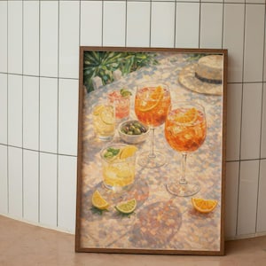 May include: A framed print depicts a still life of cocktails and appetizers. The image features several glasses filled with orange and yellow beverages, garnished with citrus slices and mint. A small bowl of olives and a straw hat are also visible.
