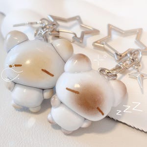 May include: Two white bear-shaped keyrings with brown accents, sleeping expressions, and silver hardware. One keyring has a star-shaped charm. The keyrings are on a white surface.