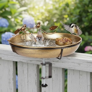 May include: A decorative bird bath with a bronze finish, mounted on a white wooden fence. Several small birds are perched on the rim and in the water. A small bowl of birdseed is also visible.