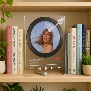 May include: A clear acrylic photo frame featuring a circular image of a woman with a light blue background. The frame has a silver-colored arm and a music player interface with the text "So High School" and "Taylor Swift."