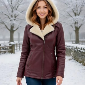 Puede incluir: Chaqueta de cuero burdeos con capucha y cuello forrados de piel sintética color crema. La chaqueta tiene cierre de cremallera y dos bolsillos delanteros. La persona lleva vaqueros azules en un entorno nevado.