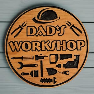 May include: A round wooden sign with the words "DAD'S WORKSHOP" and various tools carved into the wood. The sign is light brown with black detailing and a black border. A hard hat and pliers are at the top.