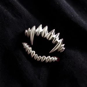 May include: A silver-colored ring shaped like a set of vampire fangs. The ring features sharp, pointed teeth and is set against a black background. The ring is a unique piece of jewelry.