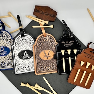 May include: An assortment of personalized golf tee holders in various colors, including blue, black, and brown. Each holder features a unique design with initials or names, and includes wooden golf tees. The holders are arranged on a dark surface.
