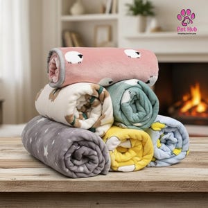 Op de afbeelding: Een stapel opgerolde huisdierendekens in verschillende kleuren en patronen. De dekens hebben ontwerpen met schapen, sterren en andere speelse motieven. De dekens zijn gerangschikt op een houten oppervlak.