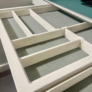 Pode incluir: Um organizador de gaveta de plástico branco com múltiplos compartimentos retangulares. O organizador foi projetado para caber dentro de uma gaveta e tem uma estética limpa e minimalista. Os compartimentos são de vários tamanhos.