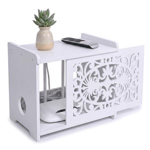May include: A white storage shelf with a decorative cutout sliding door, featuring a top surface holding a small potted succulent and a remote control, with a white router and power cables visible inside the open storage compartment.