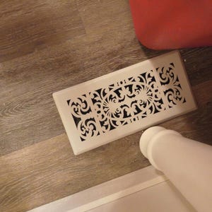 May include: A rectangular, off-white air vent cover with a decorative, openwork design. The pattern features swirling floral and geometric shapes. The vent is positioned on a wooden floor, with a red object and a white object in the background.