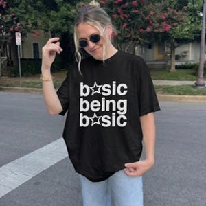 May include: A black short-sleeved t-shirt featuring white text that reads "b*sic being b*sic" with star symbols replacing the letter "a" in the word "basic". The shirt has a crew neckline and a loose, relaxed fit, made of solid black fabric.