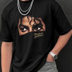 May include: A black short-sleeved t-shirt featuring a graphic print of Michael Jackson's eyes and hair with the text Michael Jackson written in a gold cursive script below the eyes. The shirt has a crew neckline and a relaxed fit design.