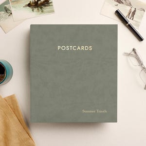 May include: A sage green rectangular postcard album with a smooth, matte cover. The word POSTCARDS is printed in gold capital letters at the top center, and the words Summer Travels are printed in smaller gold letters at the bottom right corner.
