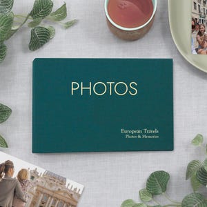 May include: A teal photo album with the word "PHOTOS" in gold lettering. Below, the text reads "European Travels Photos & Memories." The album is on a light gray surface, with a cup of tea and greenery in the background.
