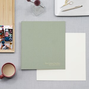 May include: A sage green hardcover book with the text "Your Custom Text Here" printed in small white letters on the bottom right corner. The book is positioned next to a blank white sheet of paper, a red mug, and a wooden tray containing a photo.